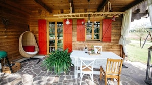 Een houten hutje met een stenen vloer, een eettafel, stoelen, een hanglamp en een groot raam met luiken.