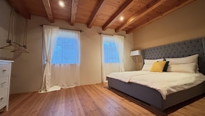 Een slaapkamer met een houten plafond, een bed met een grijs hoofdeinde en twee ramen met dunne gordijnen.