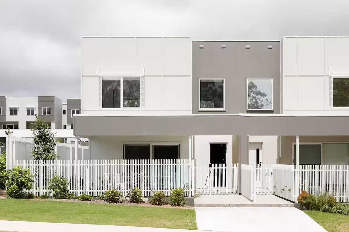 Un immeuble résidentiel moderne à deux étages, doté d’une façade blanche, de grandes fenêtres et d’une clôture blanche qui entoure la propriété.