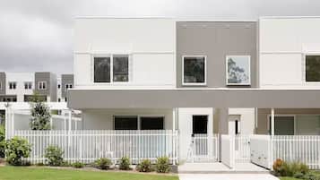 A modern two-story residential building with a white exterior, large windows, and a white fence surrounding the property.