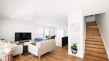 A modern living room with a flat-screen TV, a sofa, a coffee table, and a dining area with a kitchen in the background.