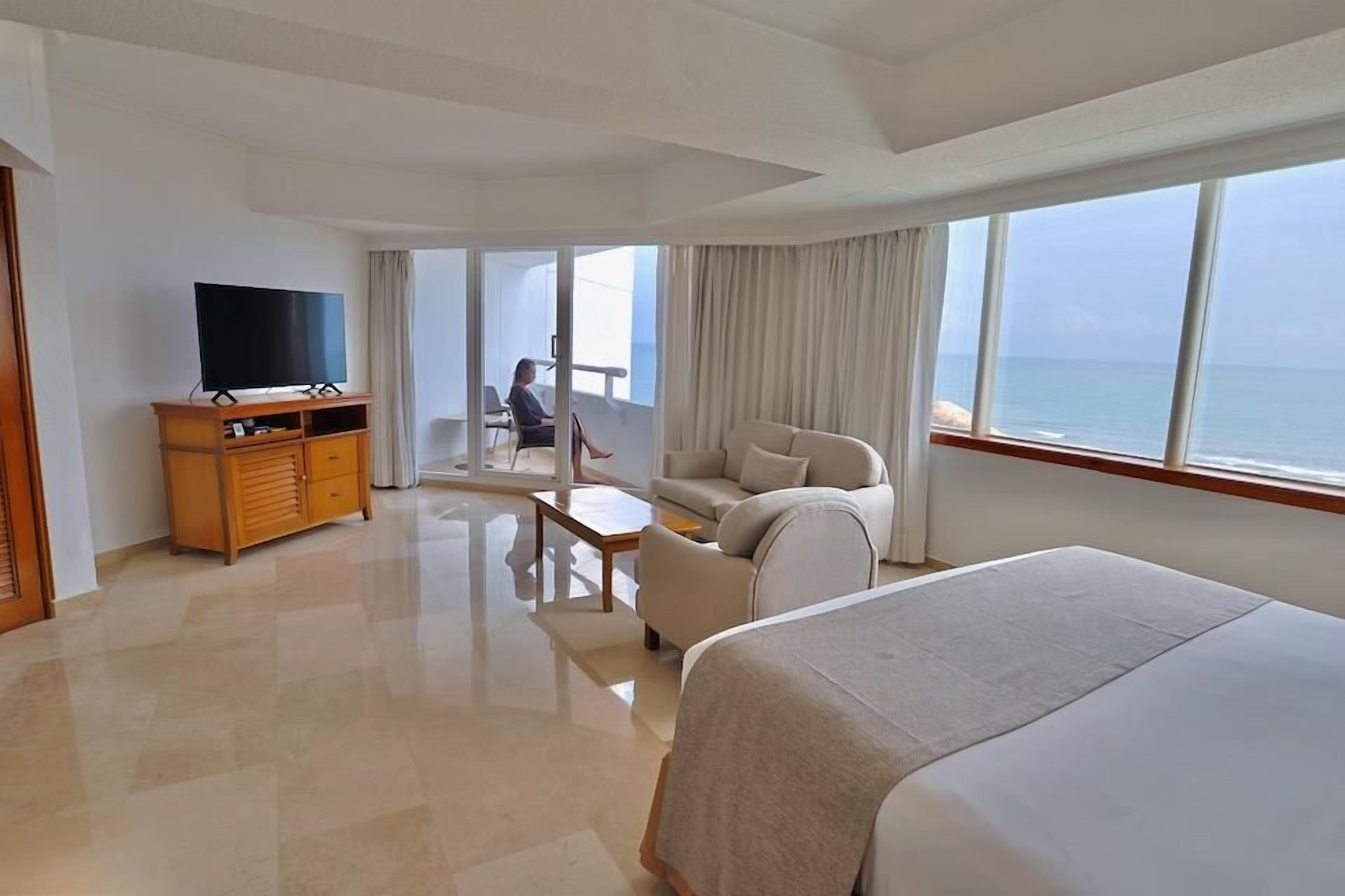 Habitació d'hotel amb llit, televisió, sofà, taula petita i balcó amb vistes al mar.