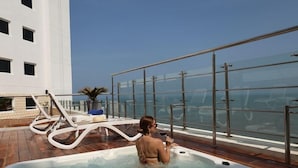 A person in a hot tub on a deck with lounge chairs and a glass railing overlooking the sea. - Hotel Almirante Cartagena Colombia (Cartagena)