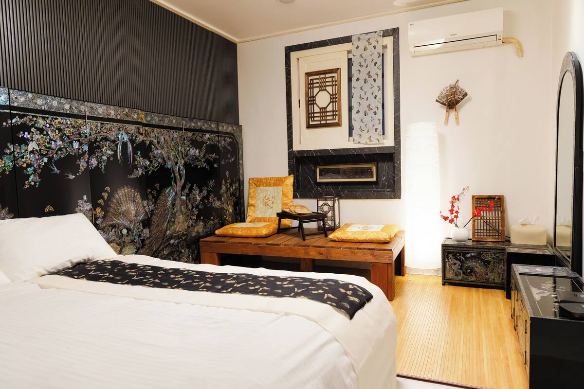 A bedroom with a bed, a wooden nightstand, a small table, a decorative wall piece, and an air conditioning unit.