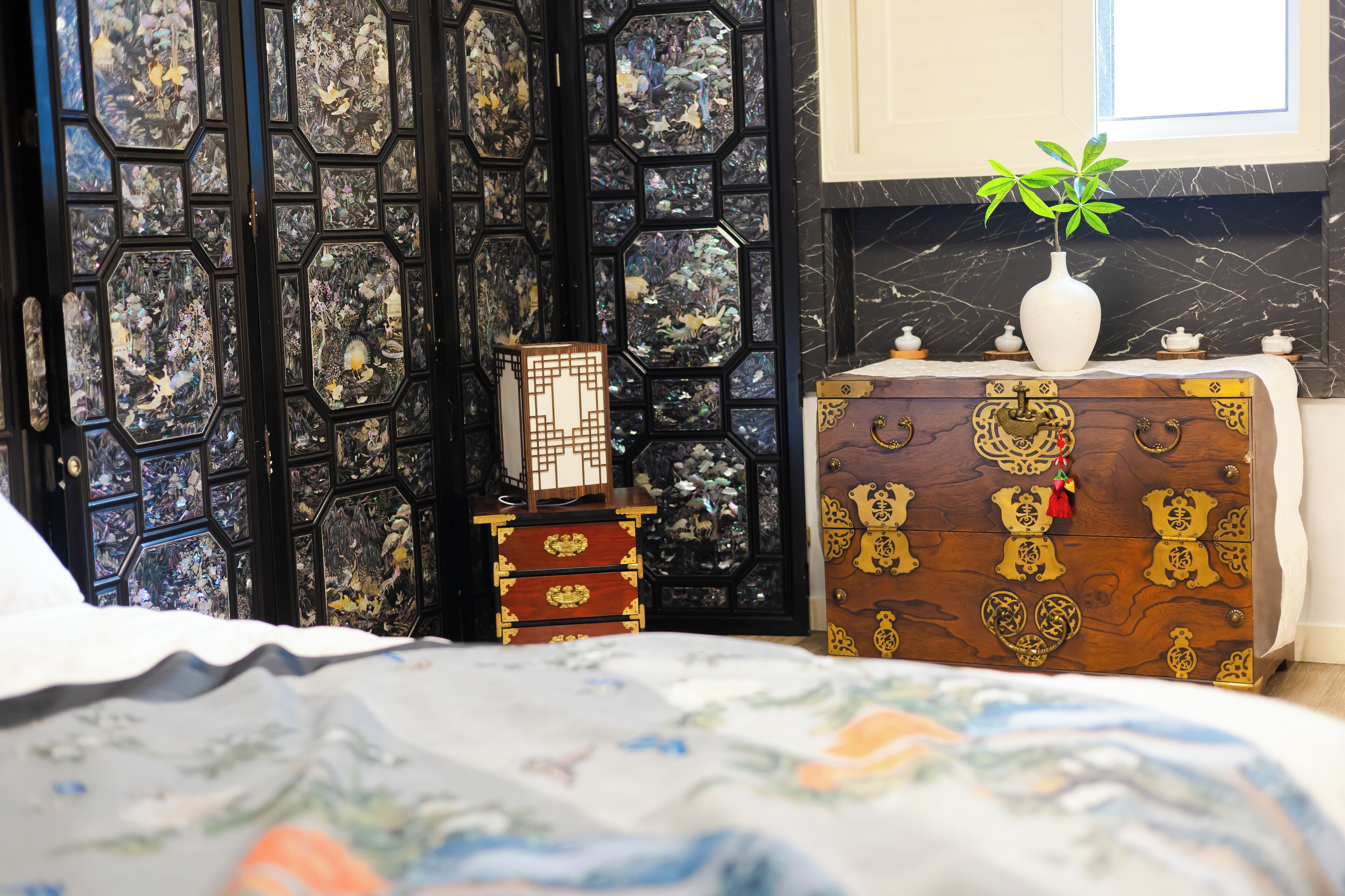 A bedroom with a bed, a dresser, a small table with a vase, and a decorative wall panel.