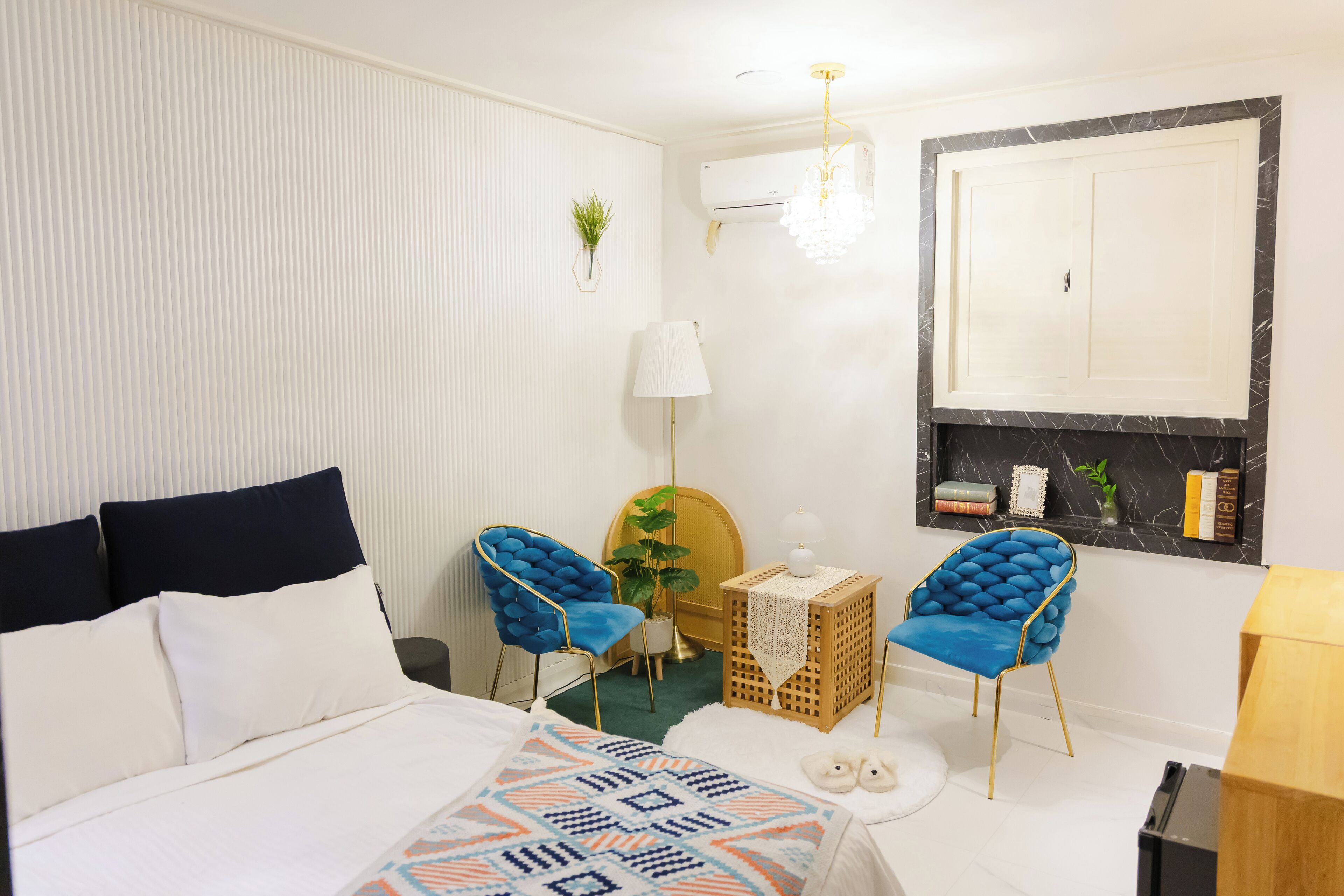 A hotel room with a bed, two blue upholstered chairs, a small wooden table, a lamp, a mirror, and a wall-mounted air conditioner.