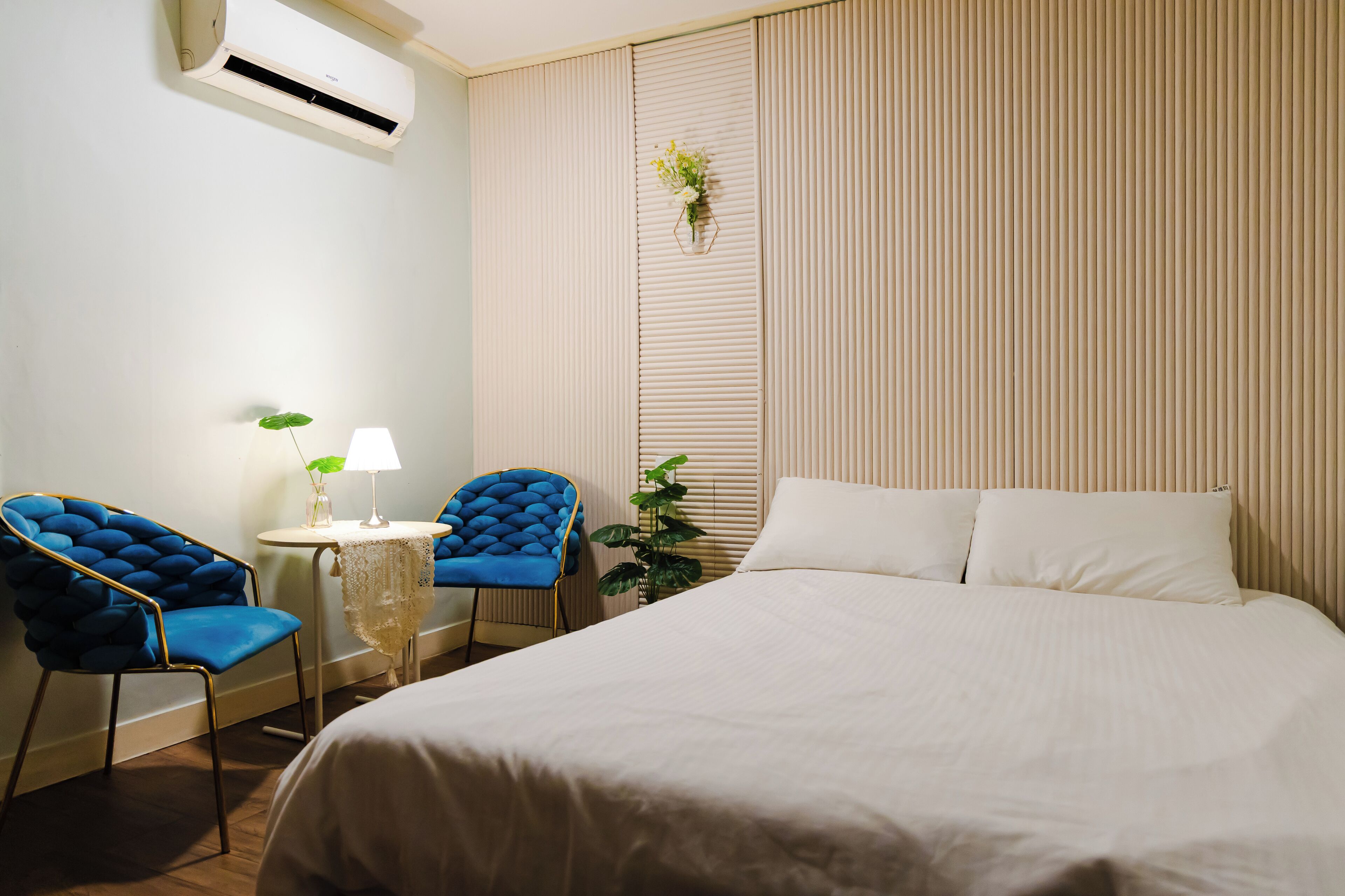 Una habitación de hotel con una cama, dos sillones azules, una mesita con una lámpara y una planta en maceta.