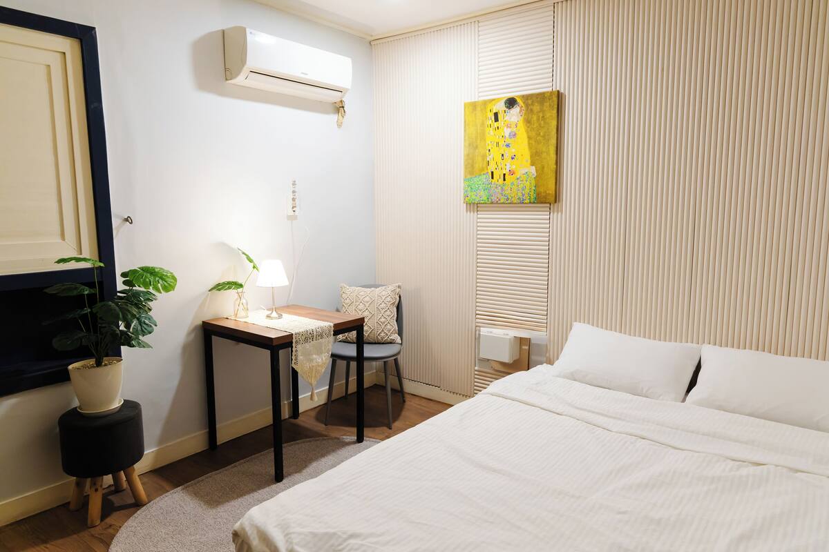 A hotel room with a bed, a desk with a lamp, a chair, a potted plant, and a painting on the wall.
