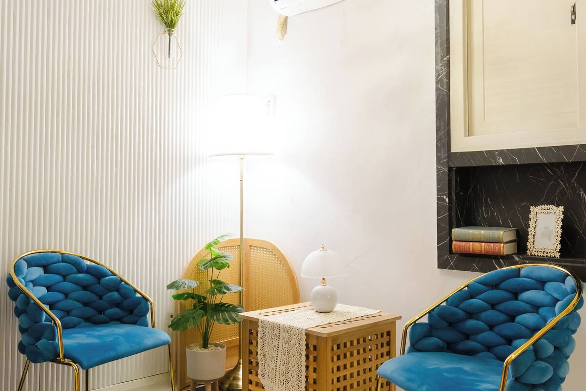 A modern room with two blue upholstered chairs, a small round table with a white lamp, a green rug, and a white fluffy rug.