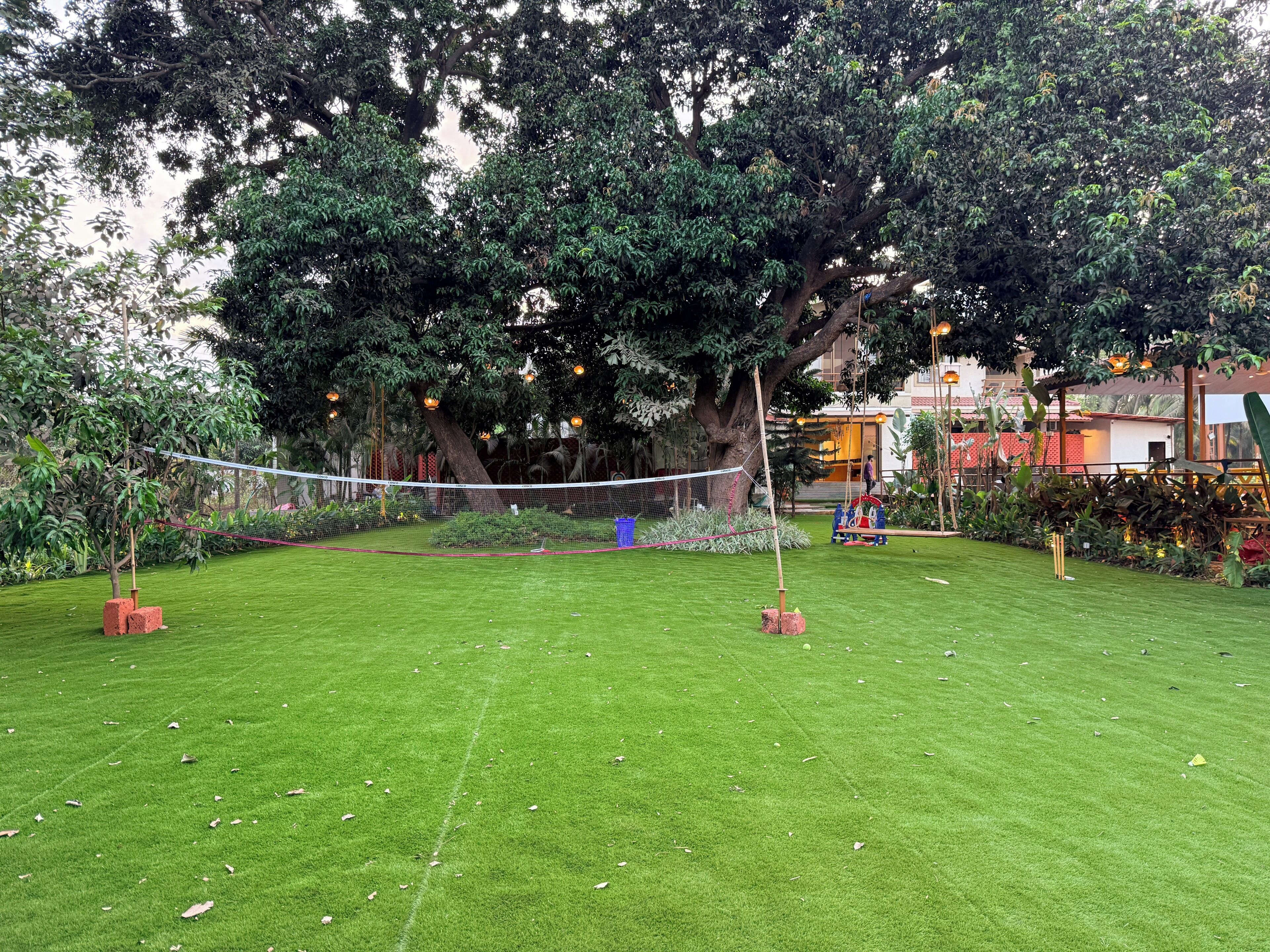 A well-maintained grassy area with a volleyball net, surrounded by trees and a building in the background.