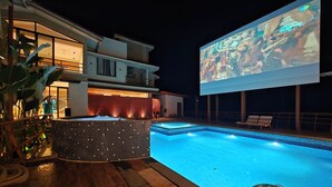 Une piscine avec un jacuzzi, des chaises longues et un grand écran extérieur.