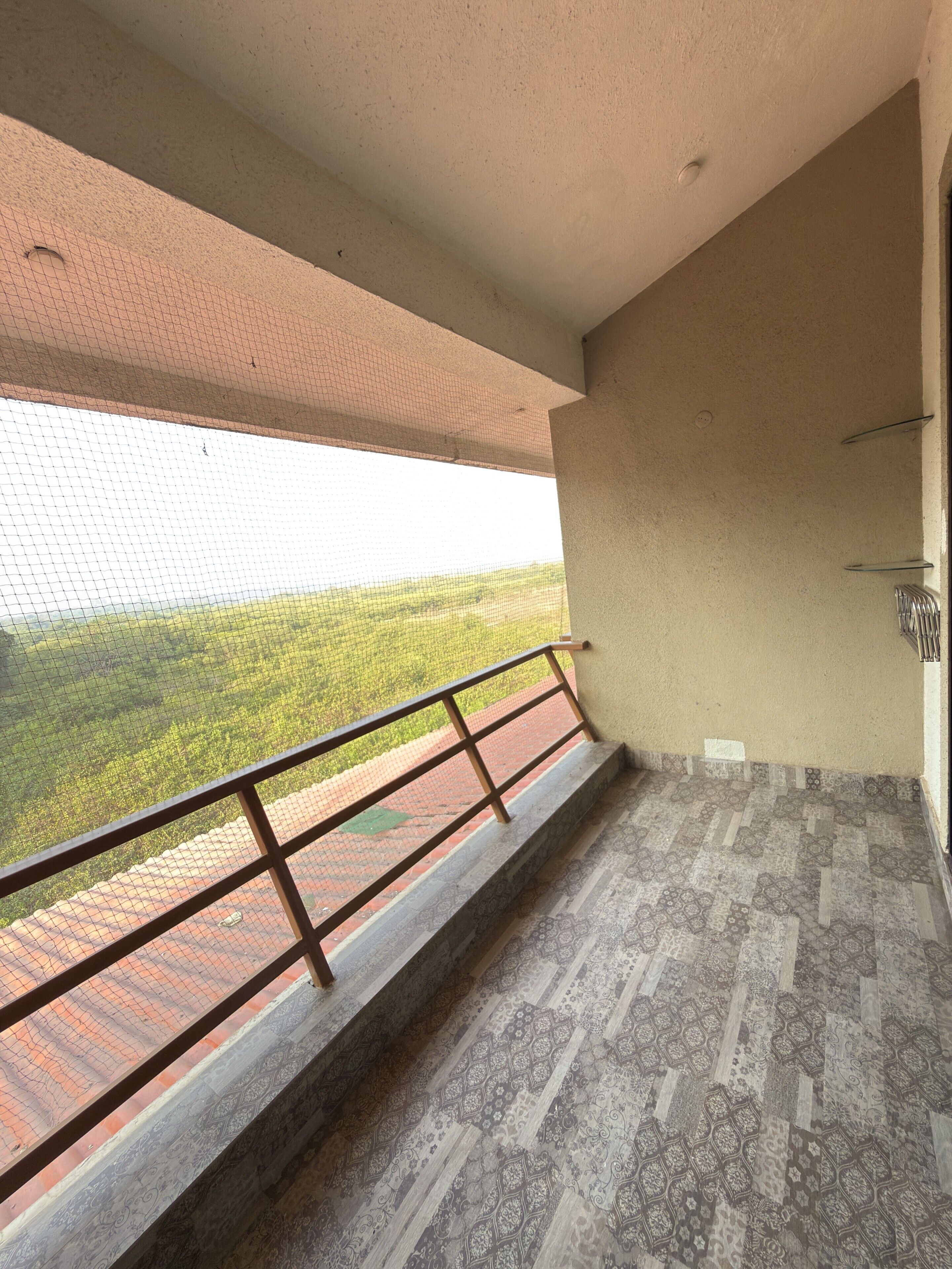 Un balcón con barandilla metálica, piso con diseño y vista a un paisaje verde a través de una malla.