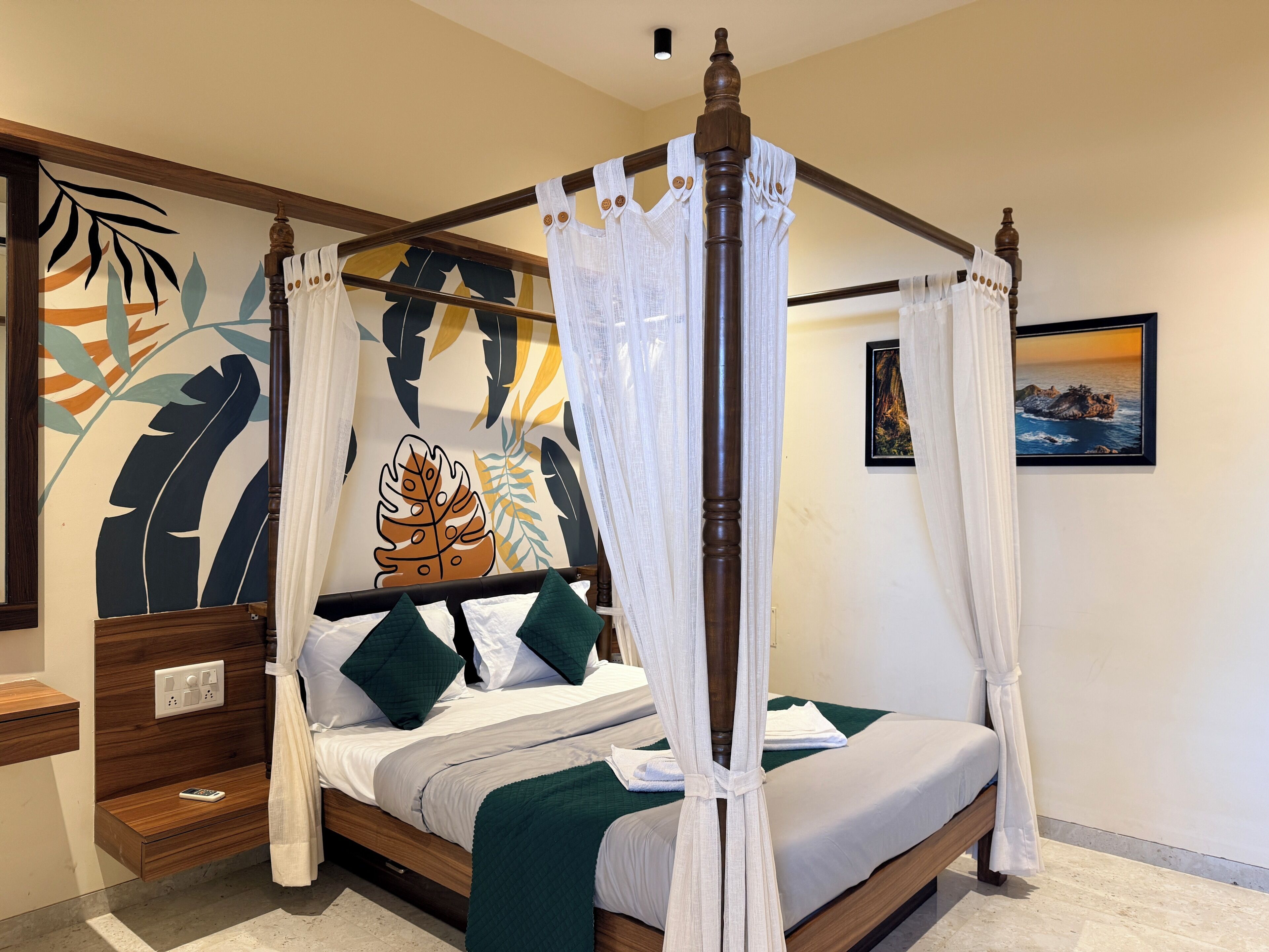 A four-poster bed with a canopy, a nightstand with a remote control, a wall with a leaf pattern, and a framed picture on the wall.