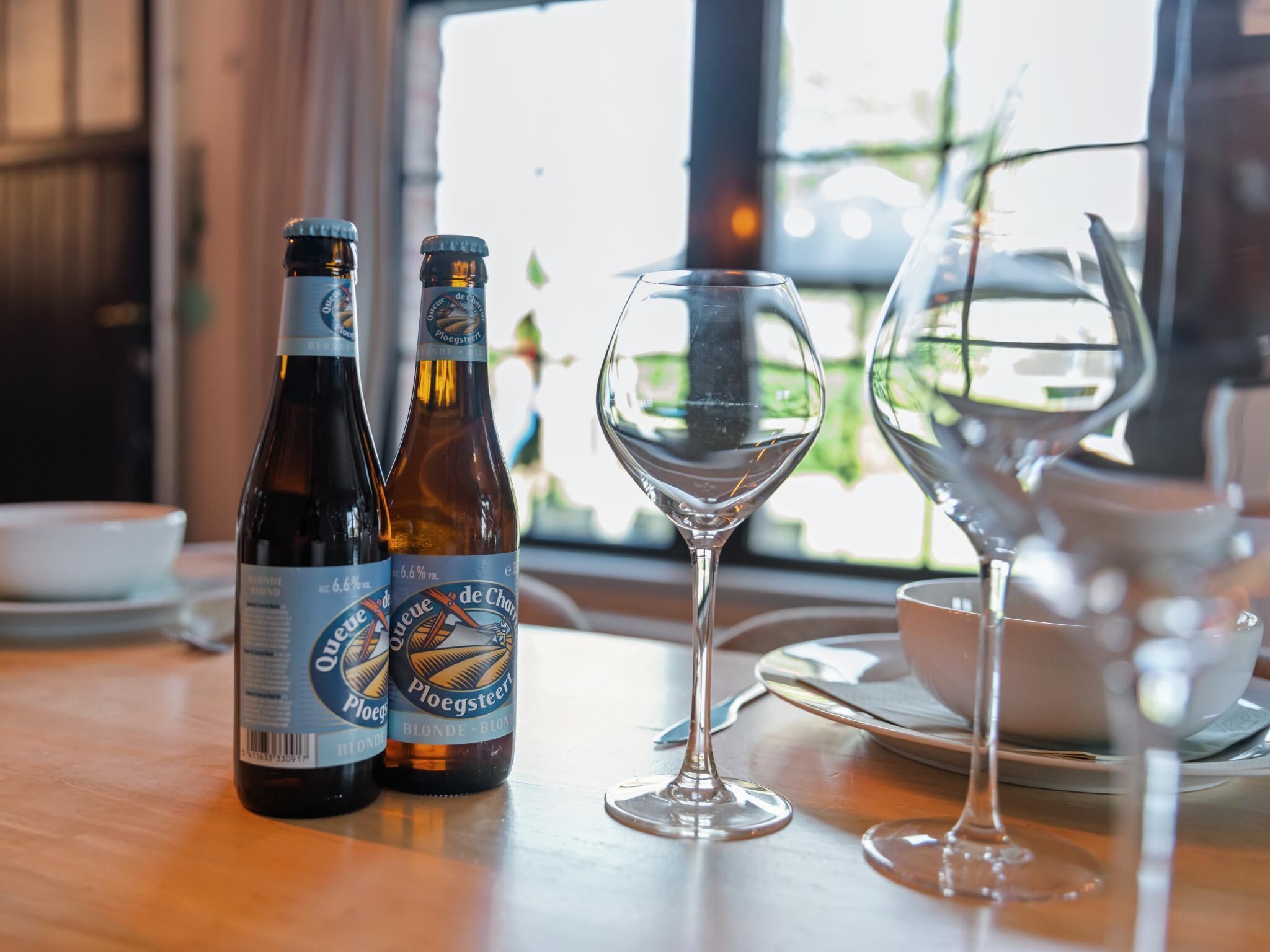 Two bottles of Blonde Blonde beer and two wine glasses on a table with a view of greenery outside.