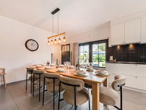 A modern dining area with a wooden table set for six, surrounded by six chairs. A hanging light fixture above the table and a wall clock to the left are visible.