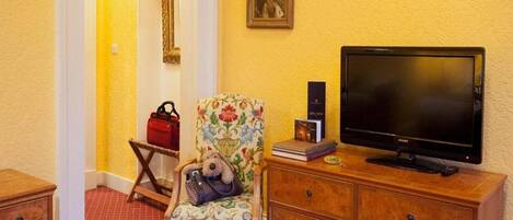 A room with a television on a wooden cabinet, a floral armchair with a handbag, a red handbag, and a framed picture on the wall.