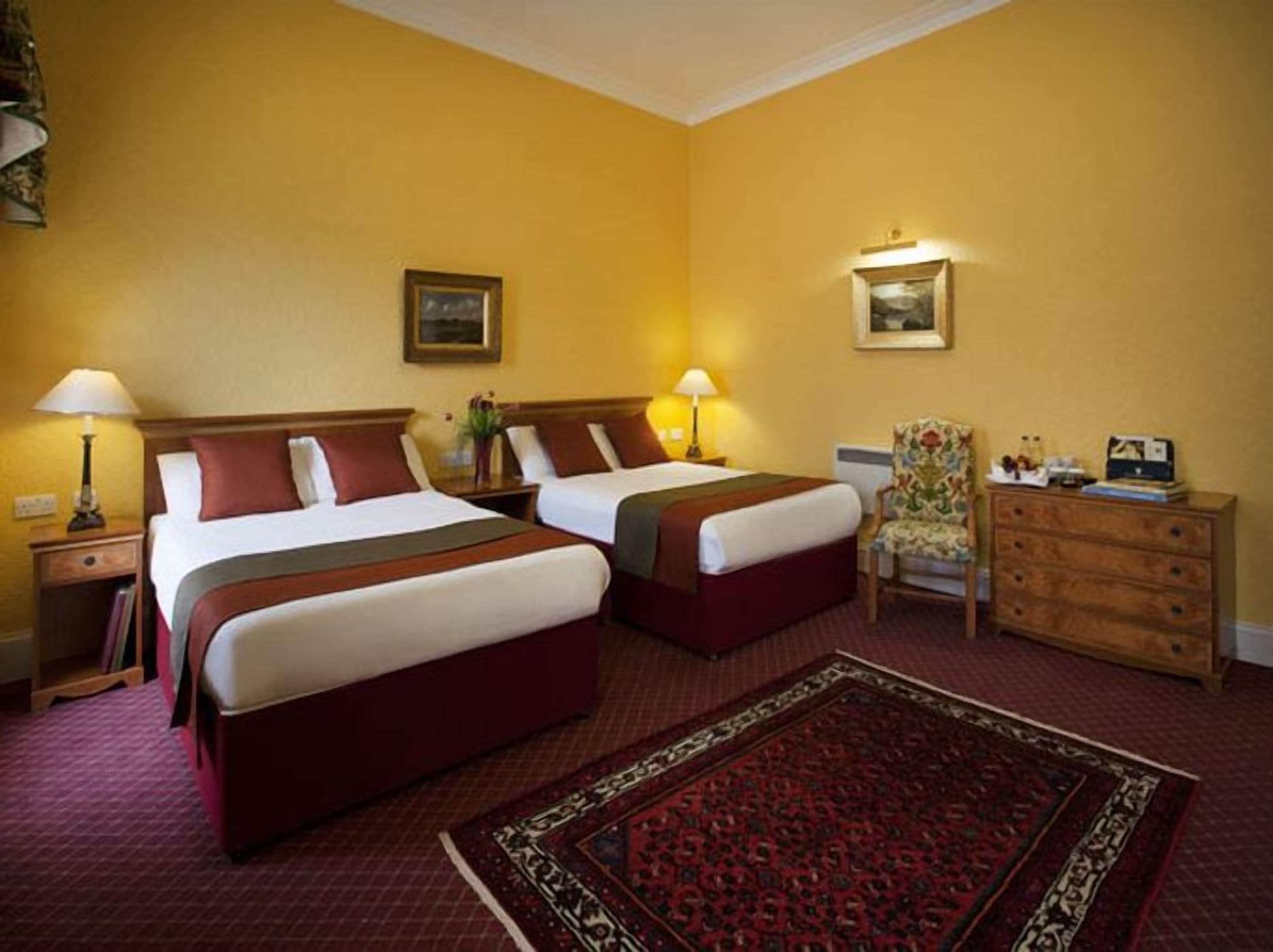 Hotellihuone, jossa on kaksi sänkyä, puinen lipasto, tuoli ja matto.