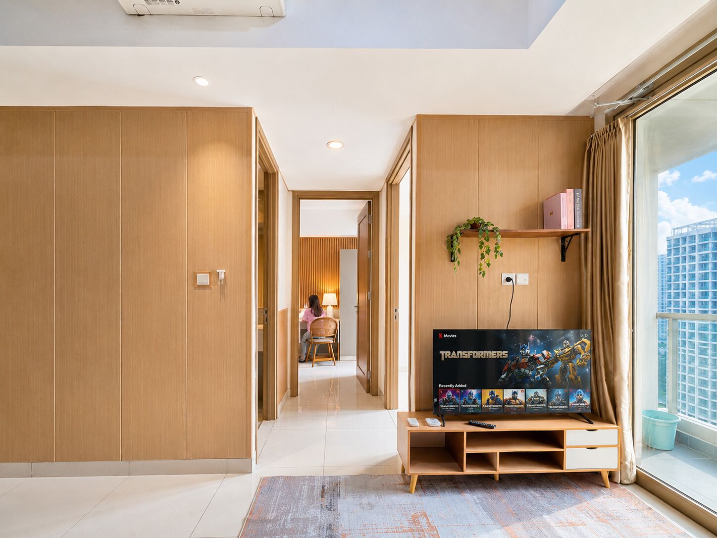 Une chambre d’hôtel moderne dotée d’une grande télévision, de lambris en bois, d’une vue sur la ville et d’un balcon agrémenté d’une plante.