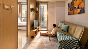 A modern living room with a sofa, a coffee table, a TV, and a large window with a city view. - Alia By SanLiving - 2BR - HubLife Mall (Jakarta)