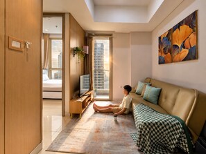 A modern living room with a sofa, a coffee table, a TV, and a large window with a city view. - Alia By SanLiving - 2BR - HubLife Mall (Jakarta)