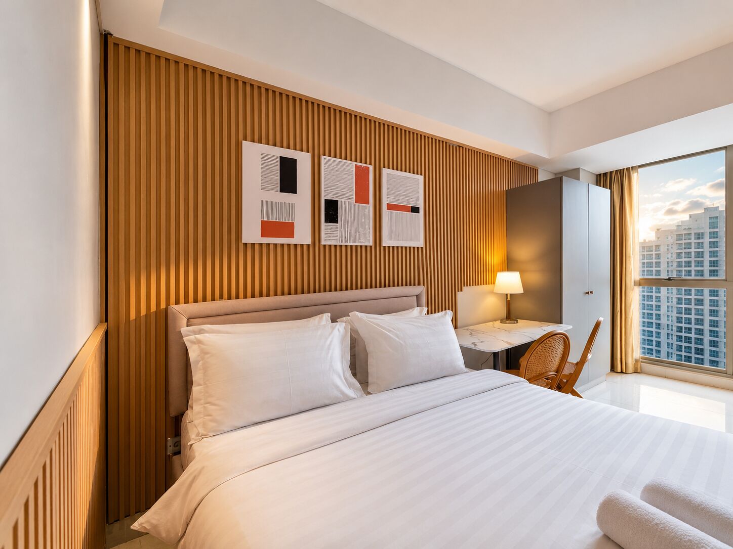Una camera d'albergo con un letto, una scrivania con una lampada, una sedia e vista sulla città attraverso ampie finestre.