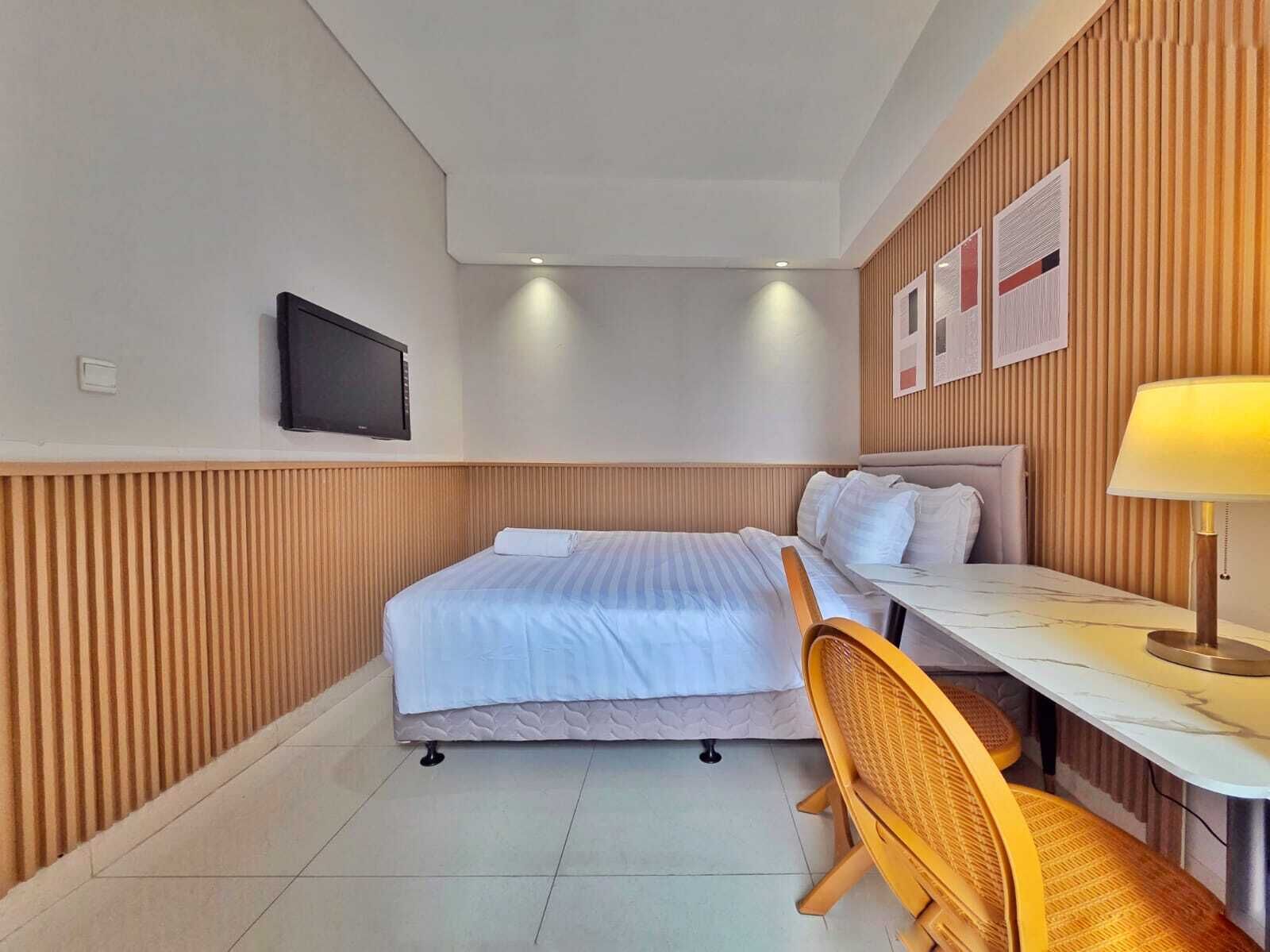 A hotel room with a bed, a desk, a chair, a TV, and a wall-mounted lamp.