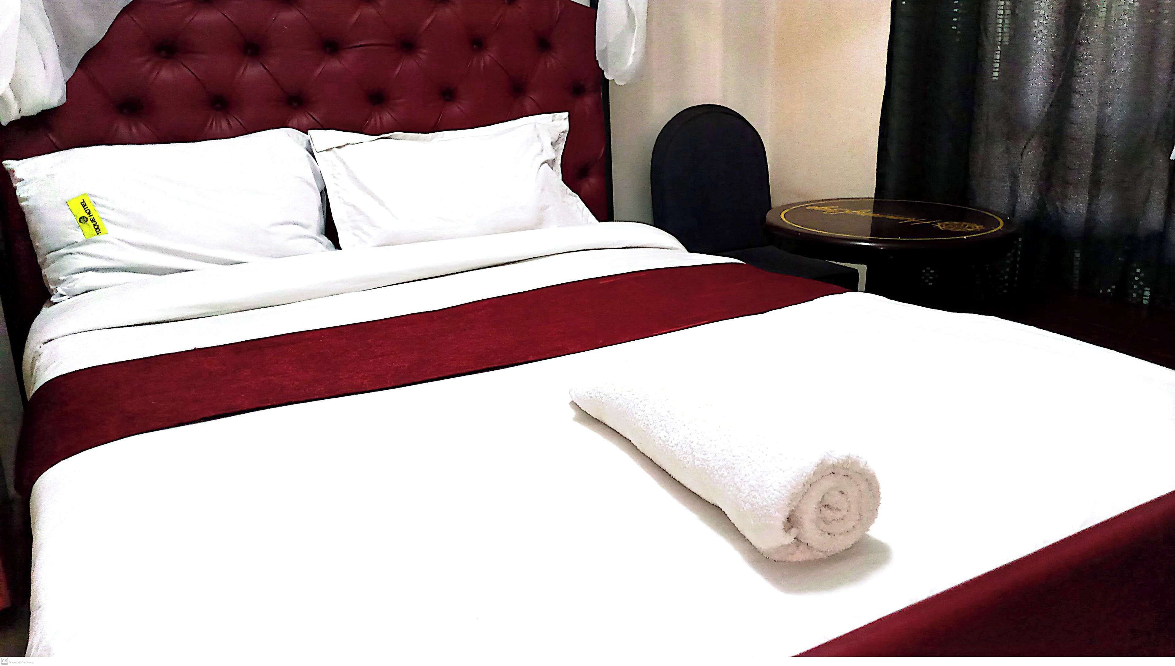 Een bed met een rood hoofdeinde, wit beddengoed en een opgerolde witte handdoek op het bed.