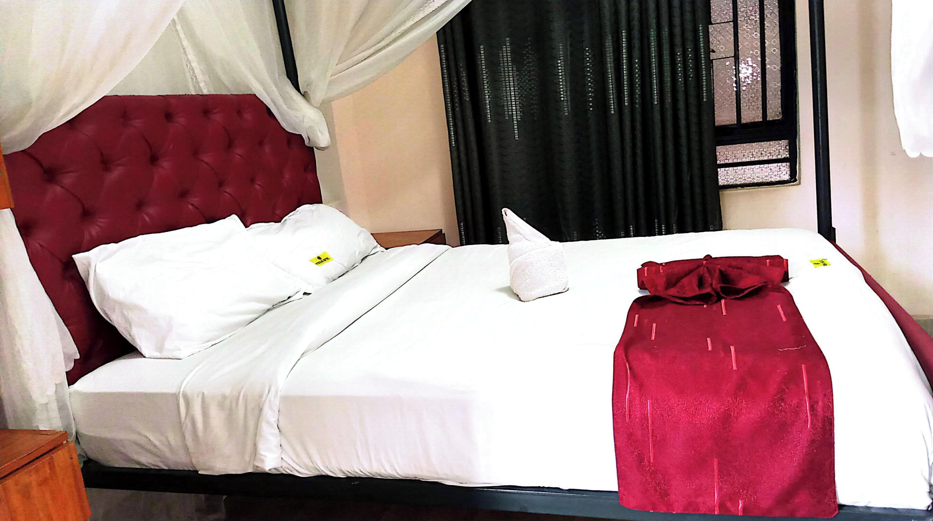 A neatly made bed with white linens and a red blanket, a tufted headboard, and a window with black curtains.