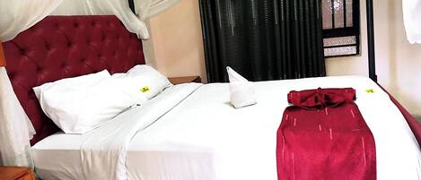 A neatly made bed with white linens and a red blanket, a tufted headboard, and a window with black curtains.