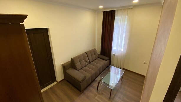 Ein Zimmer mit braunem Sofa, einem Glastisch, einer Holztür und einem Fenster mit Vorhängen.