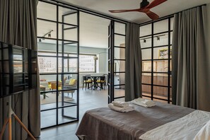 A modern hotel room with a large bed, a ceiling fan, and a view of the city through floor-to-ceiling windows. - Luxurious hideaway loft at Psyri (Athina)
