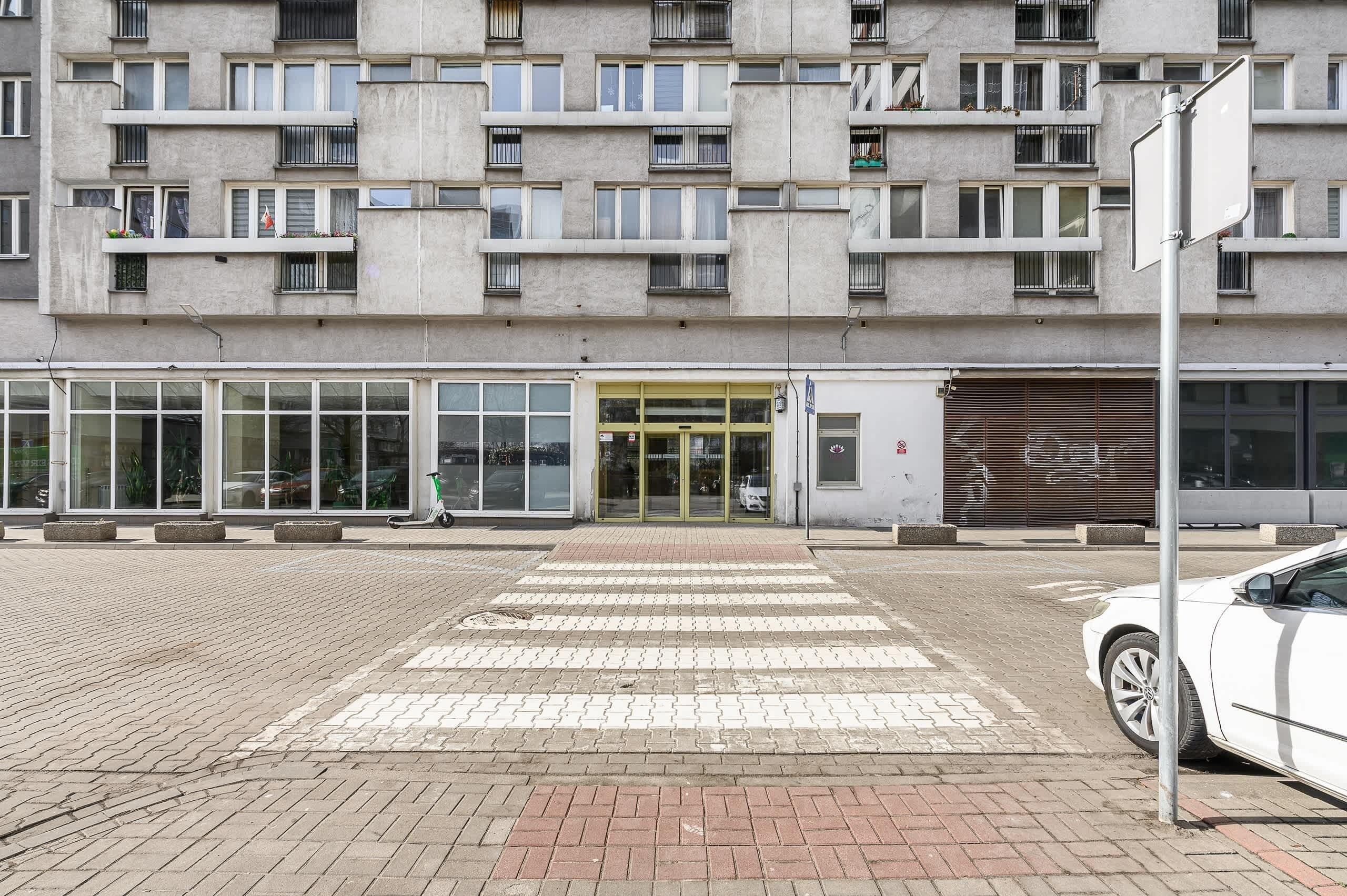 Un immeuble à étages doté d’un style architectural moderne, avec une grande entrée en verre et un espace de stationnement où se trouve une voiture blanche.
