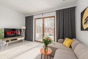 A modern living room with a flat-screen TV, a sofa, a coffee table with a vase of flowers, and a sliding glass door to the outside. - Lumina Family Apartment patio with private garden (New apartment)