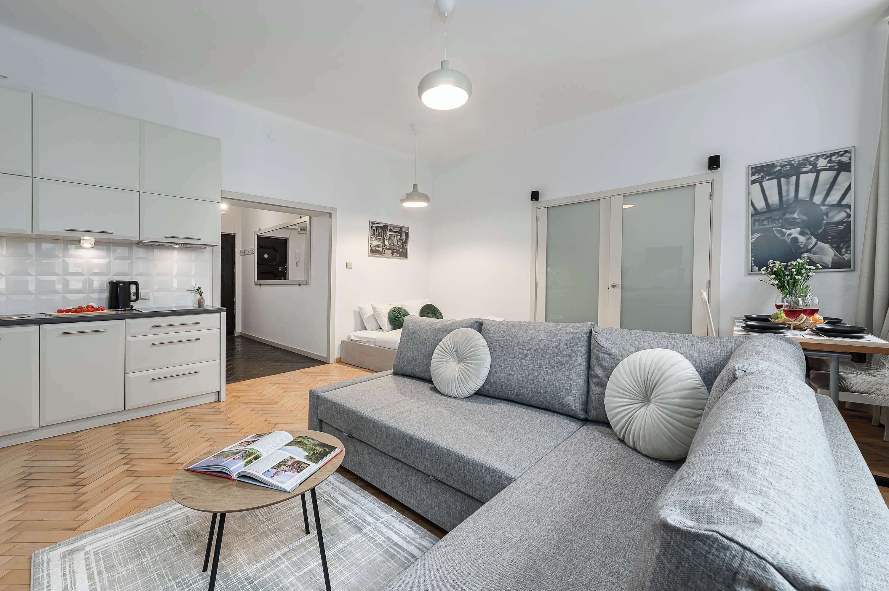 Una sala de estar moderna con un sofá gris, una mesita de centro pequeña y una zona de cocina con armarios blancos y un suelo con patrón de espiga.
