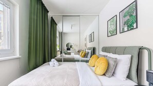 Un lit bien fait, avec une literie blanche et deux oreillers, un rideau vert et un miroir qui reflète la pièce.
