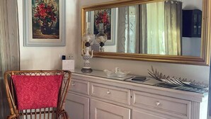 Ein Zimmer mit einem Korbstuhl, einem weißen Schminktisch mit Spiegel und einem Gemälde an der Wand.