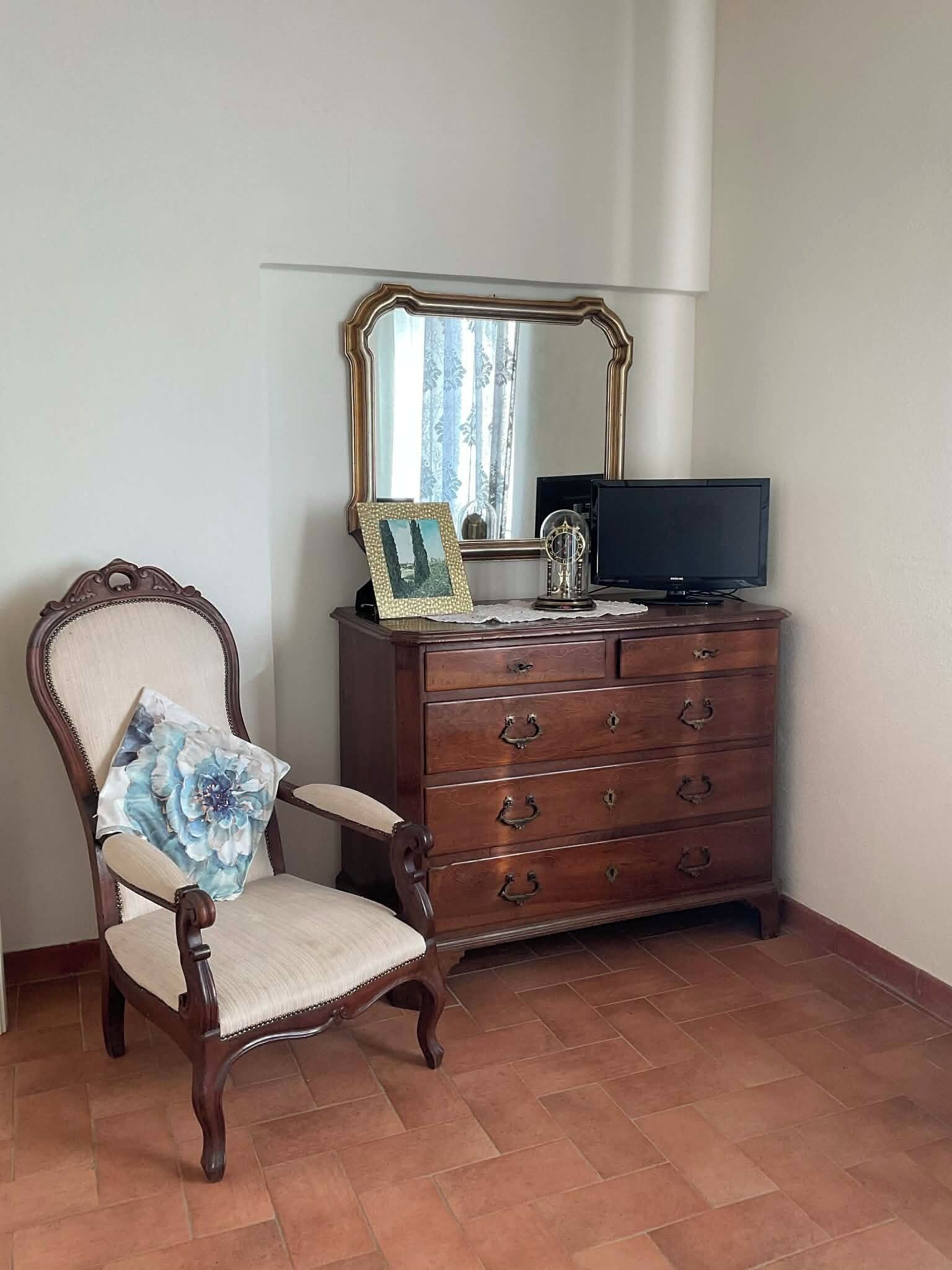 Une pièce comprenant une chaise en bois, une commode avec des tiroirs, un miroir et un téléviseur.