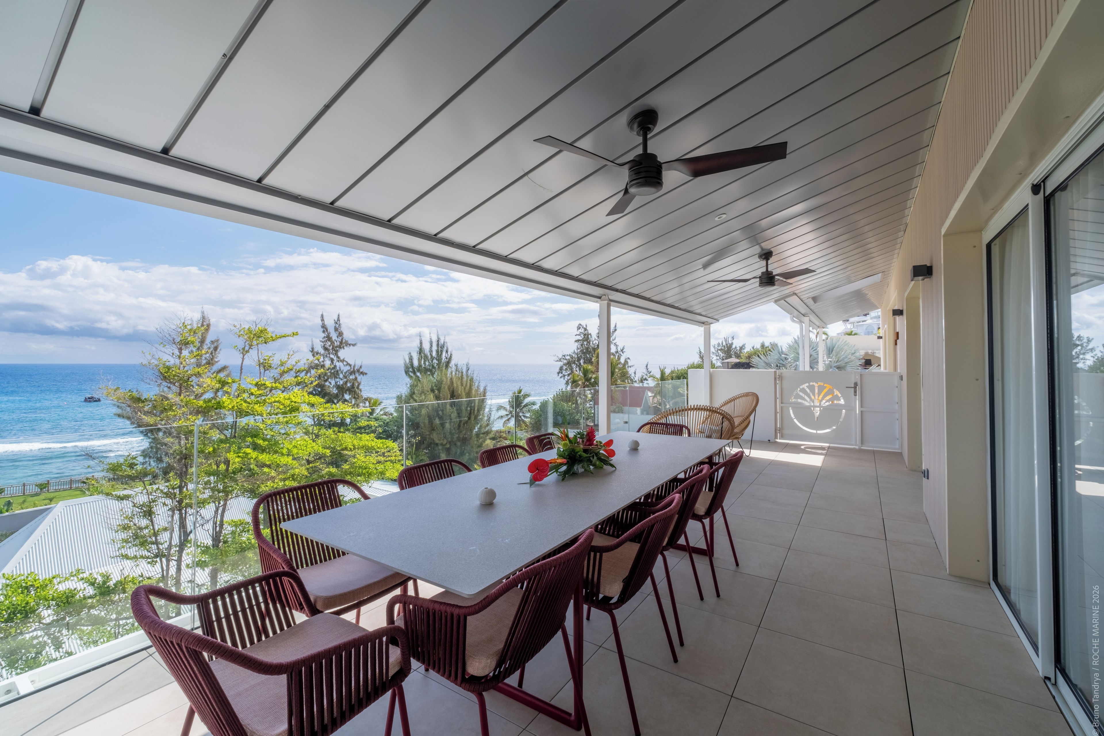 A covered outdoor dining area with a view of the sea, featuring a table with chairs and a ceiling fan.