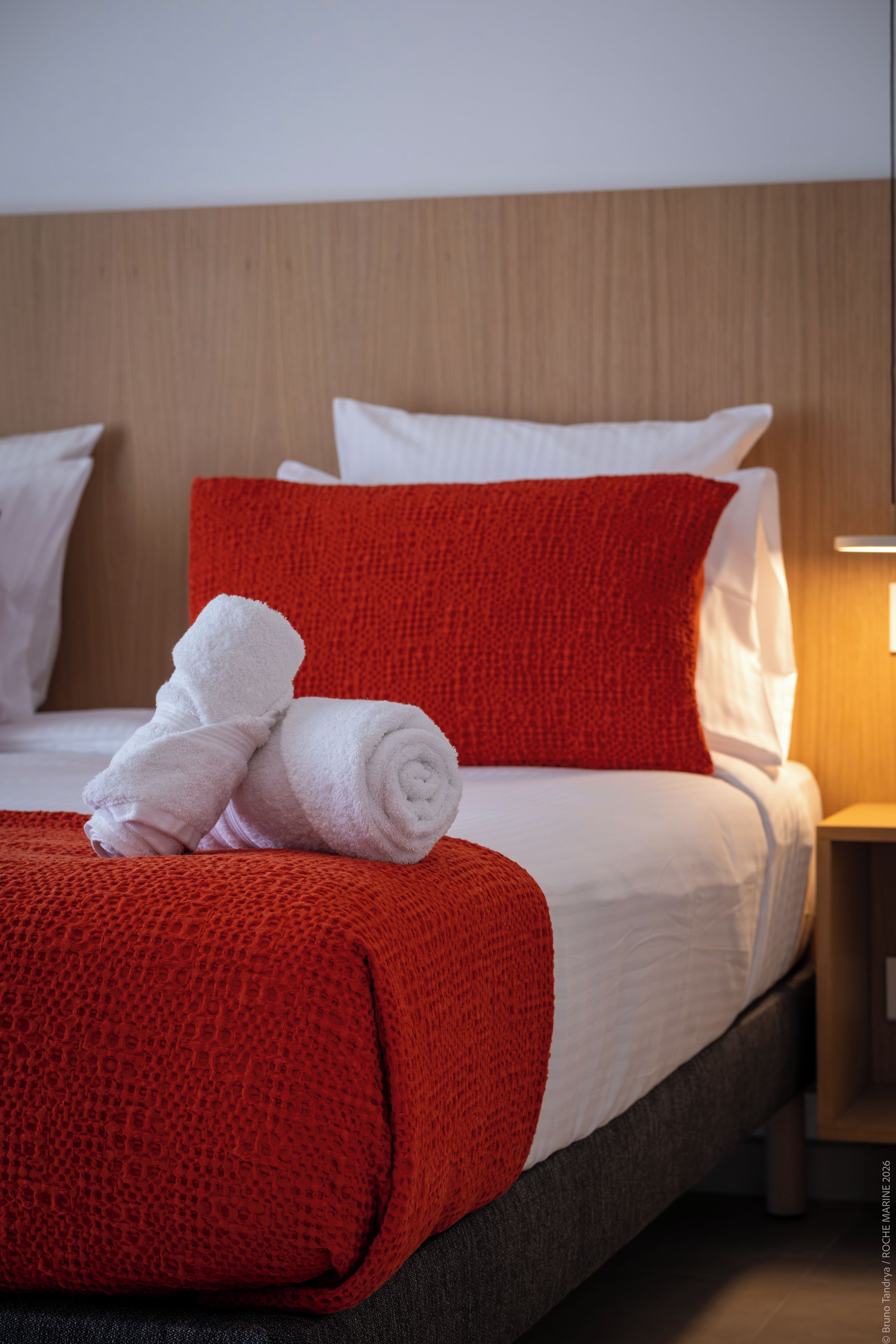 A hotel room with a neatly made bed, a red throw blanket, and white towels.