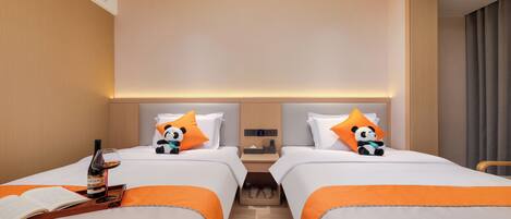 A hotel room with two beds, each with an orange accent, a bedside table with a bottle and glasses, and a nightstand with a lamp.