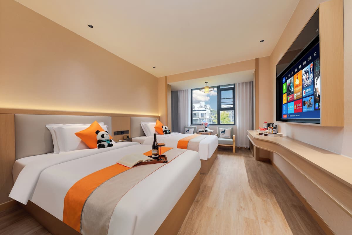 Ein Hotelzimmer mit zwei Betten, einem an der Wand befestigten Fernseher und einem Fenster mit Blick auf Gebäude.