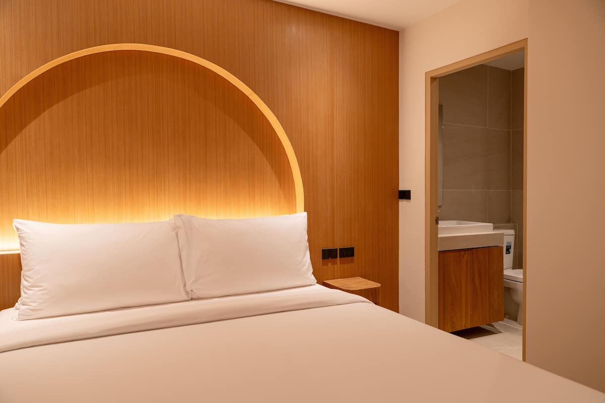 A modern hotel room with a curved wooden headboard, a white bed, a wooden nightstand, and a bathroom with a white sink and toilet.