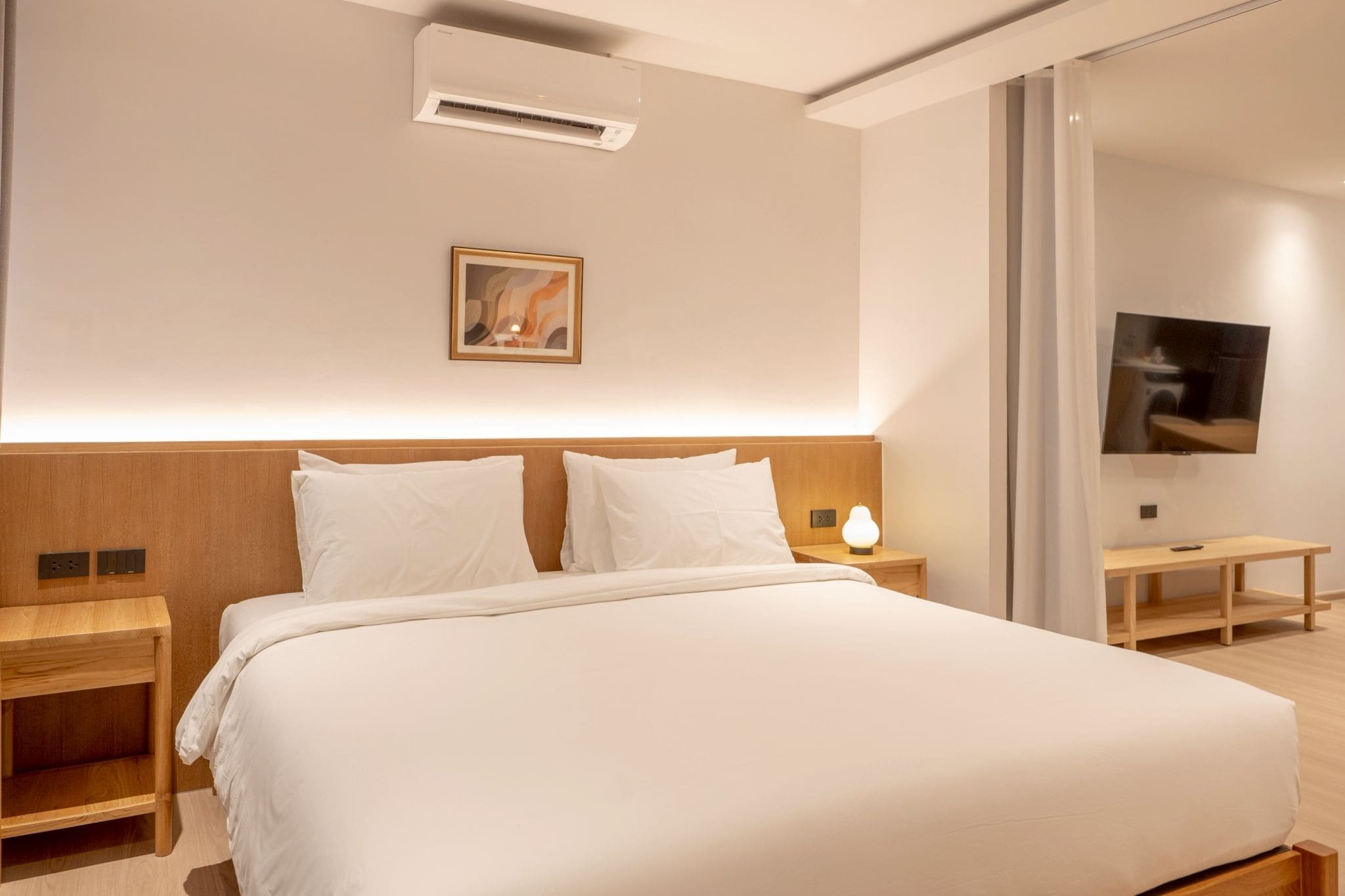 A modern hotel room with a large bed, wooden headboard, bedside tables, a flat-screen TV, and a painting on the wall.