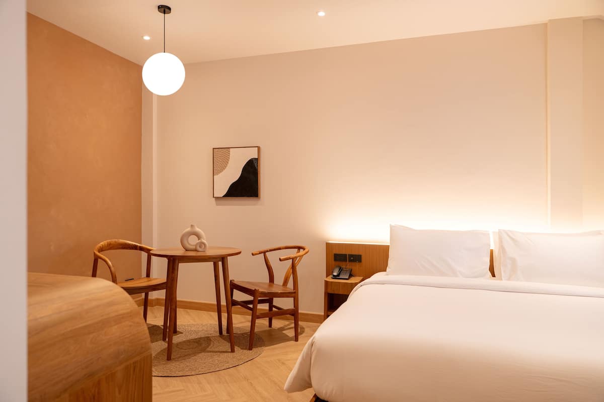 A hotel room with a bed, a desk, two chairs, a small table, a wall-mounted phone, and a wall art piece.