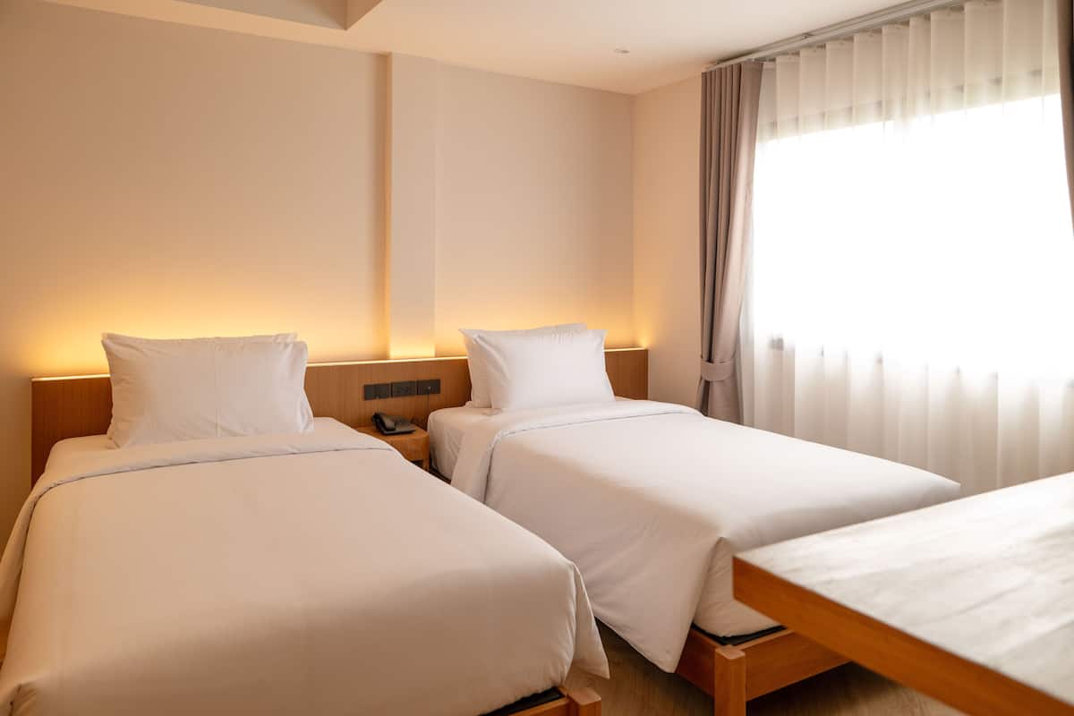 A hotel room with two beds, a wooden headboard, a bedside table with a phone, and a window with curtains.