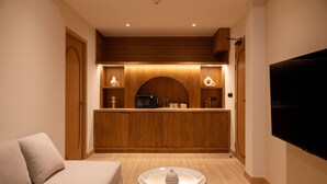 A modern hotel room with a sofa, a dining table with a white centerpiece, a kitchen area with wooden cabinets, and a flat-screen TV mounted on the wall. - Taupe Hotel (Chiang Mai)