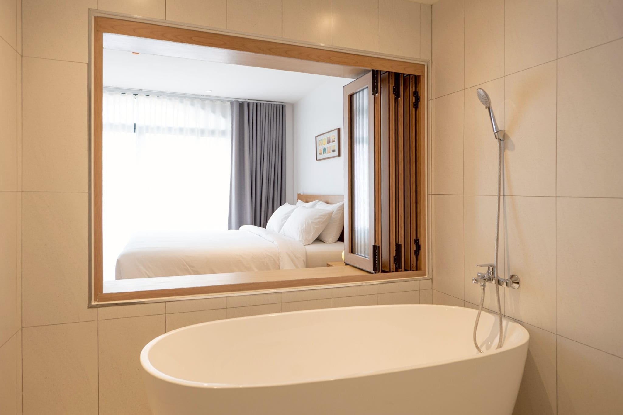 A modern bathroom with a large bathtub, a shower, and a mirror reflecting a bedroom with a bed and curtains.