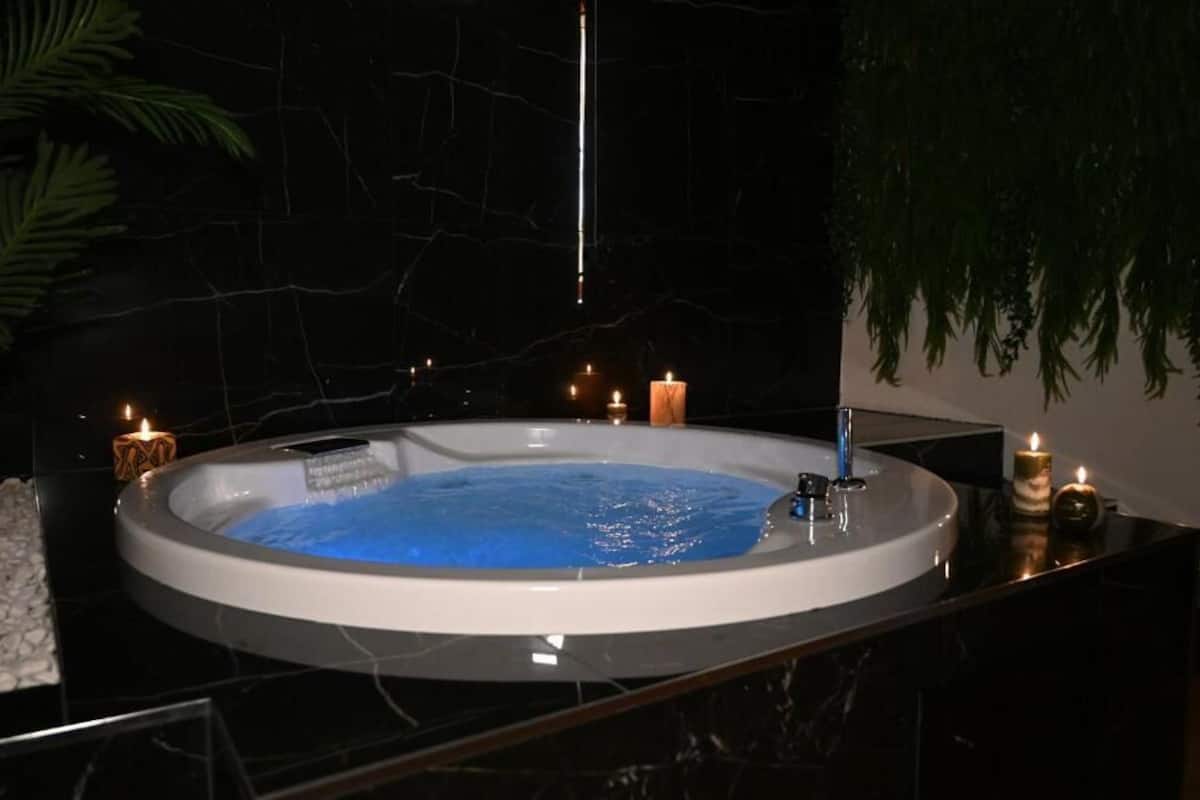 Een hottub met een blauw interieur, omringd door witte kiezelstenen, kaarsen en een donkere marmeren muur.