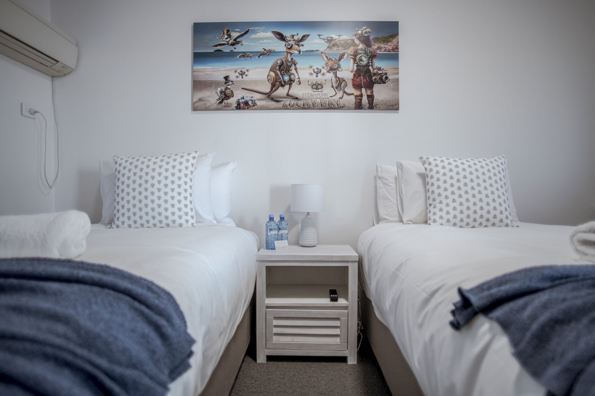 Ett hotellrum med två sängar, ett nattduksbord med en lampa, vattenflaskor och en tavla på väggen.