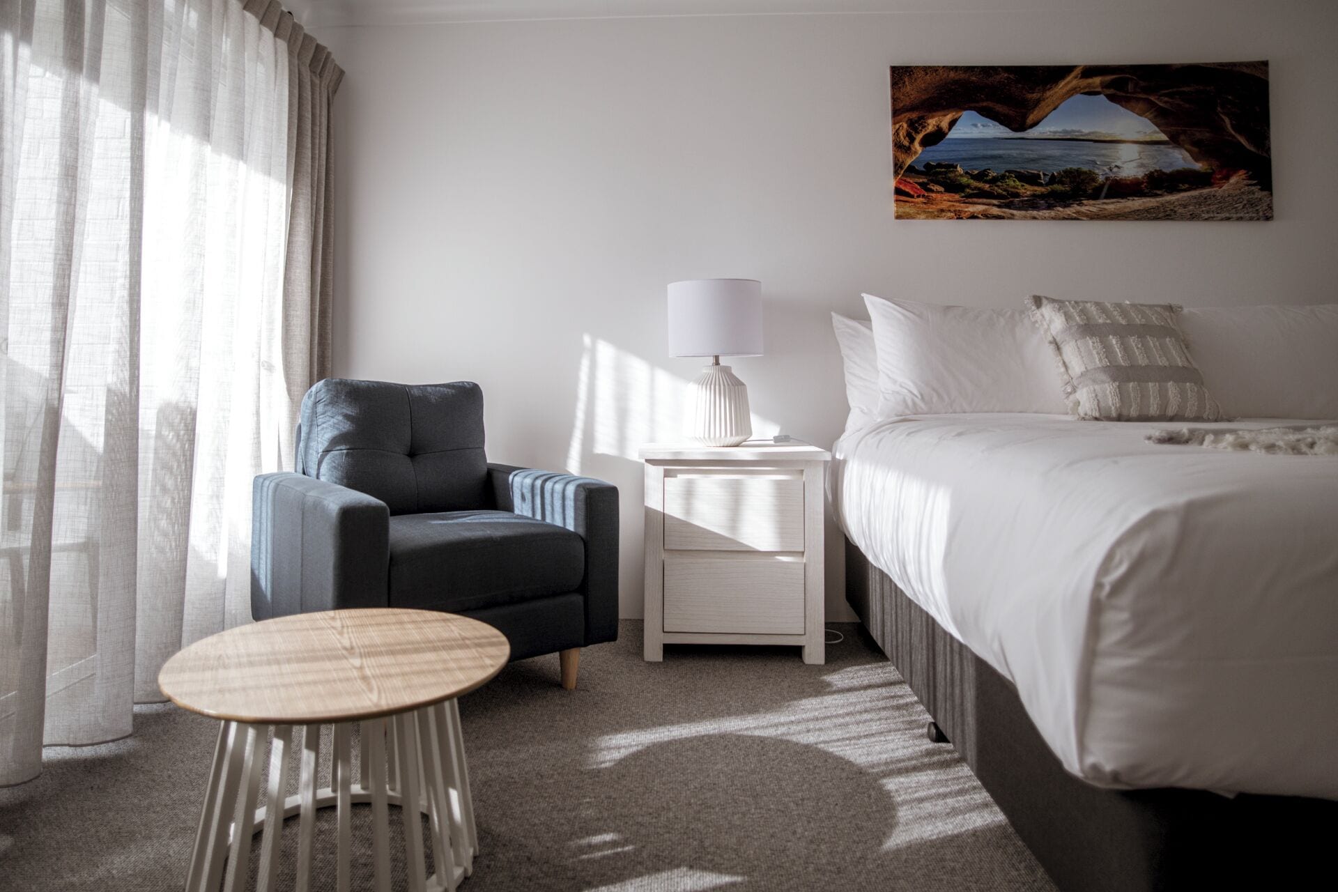 A hotel room with a bed, a chair, a small table, a nightstand with a lamp, and a painting on the wall.